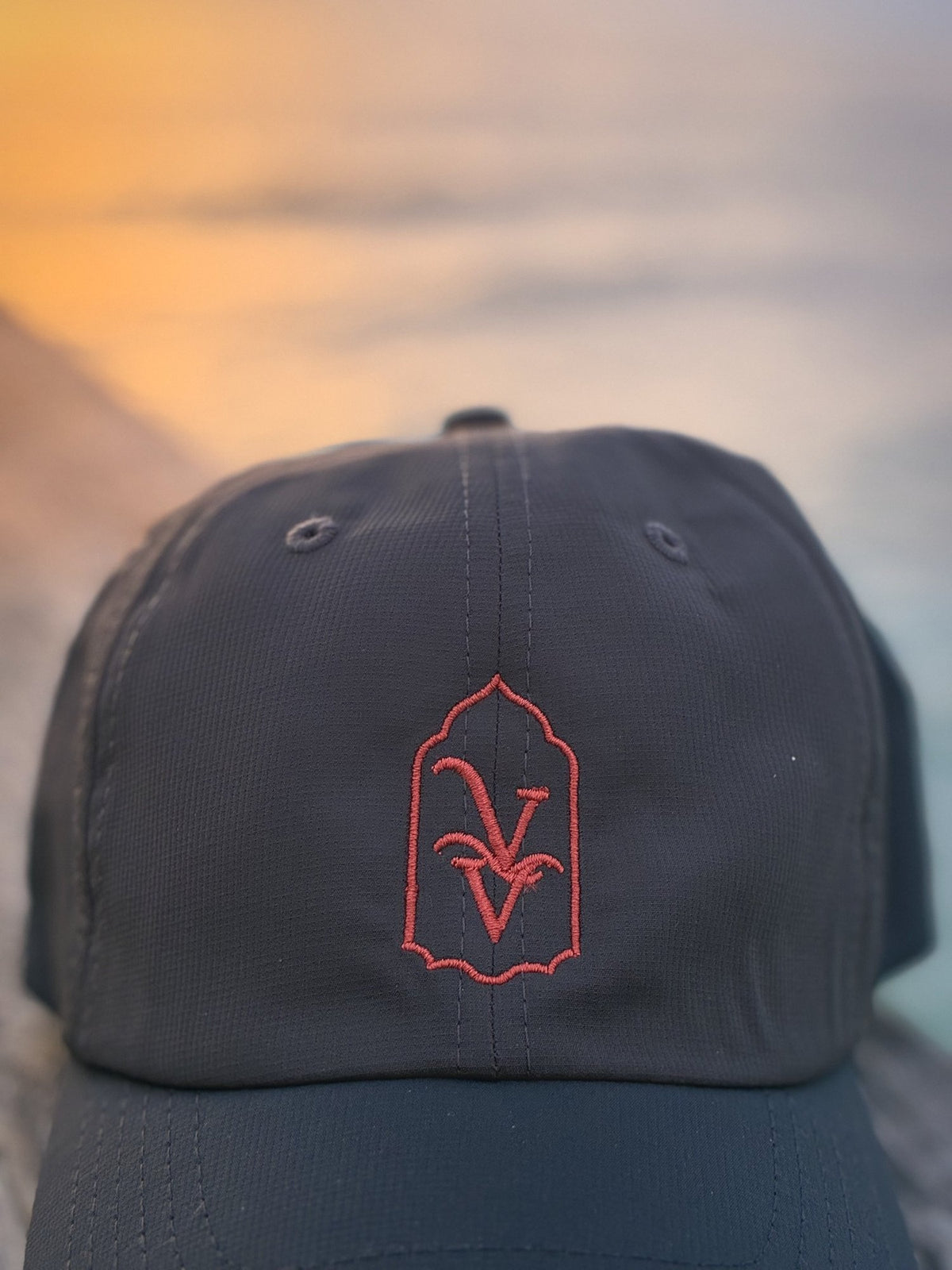 Close-up of the coral VV embroidery on the Venice Vida Afterglow Hat in Dark Grey against a golden sunset
