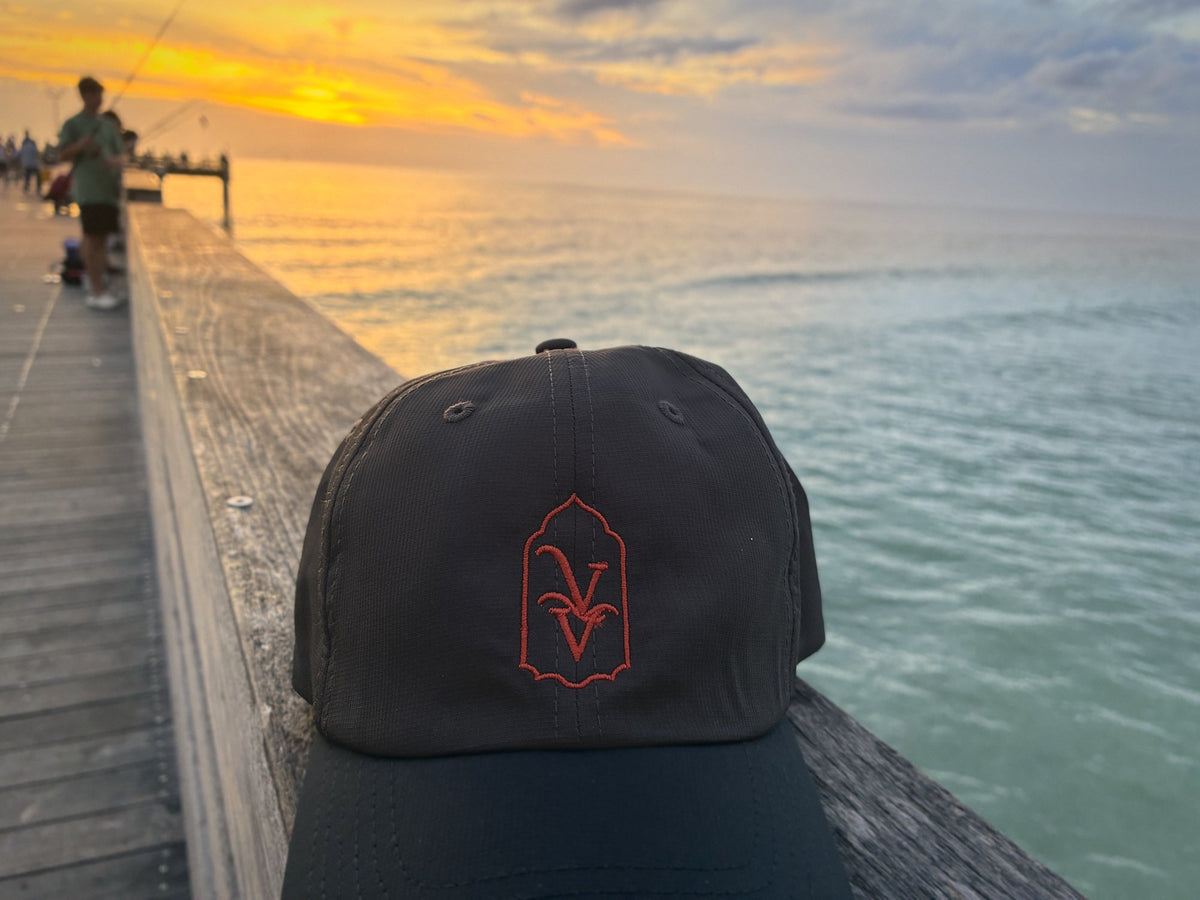 Venice Vida Afterglow Hat in Dark Grey with coral embroidered VV mark on the Venice Fishing Pier at sunset over the Gulf