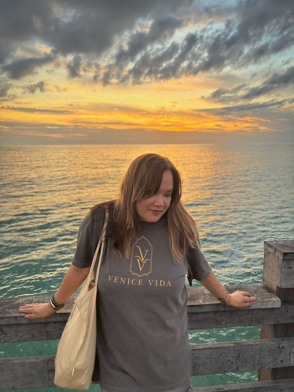 Venice Vida co-founder wearing the Afterglow Statement Tee with gold VV mark on the Venice Fishing Pier at sunset over the Gulf