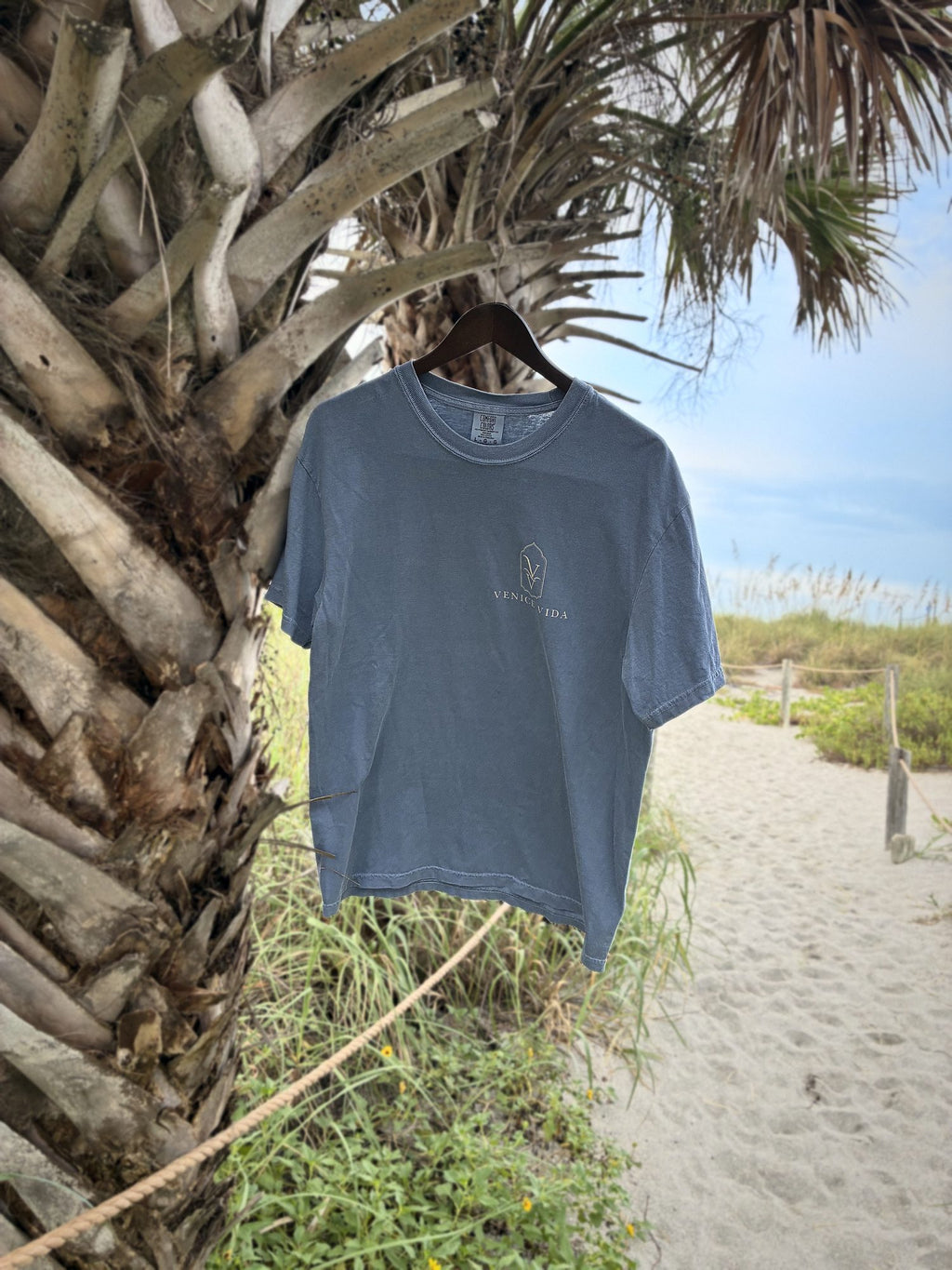 Venice Vida Signature Gulf Tee in Adriatic Blue hanging from a palm tree on the beach path in Venice, Florida