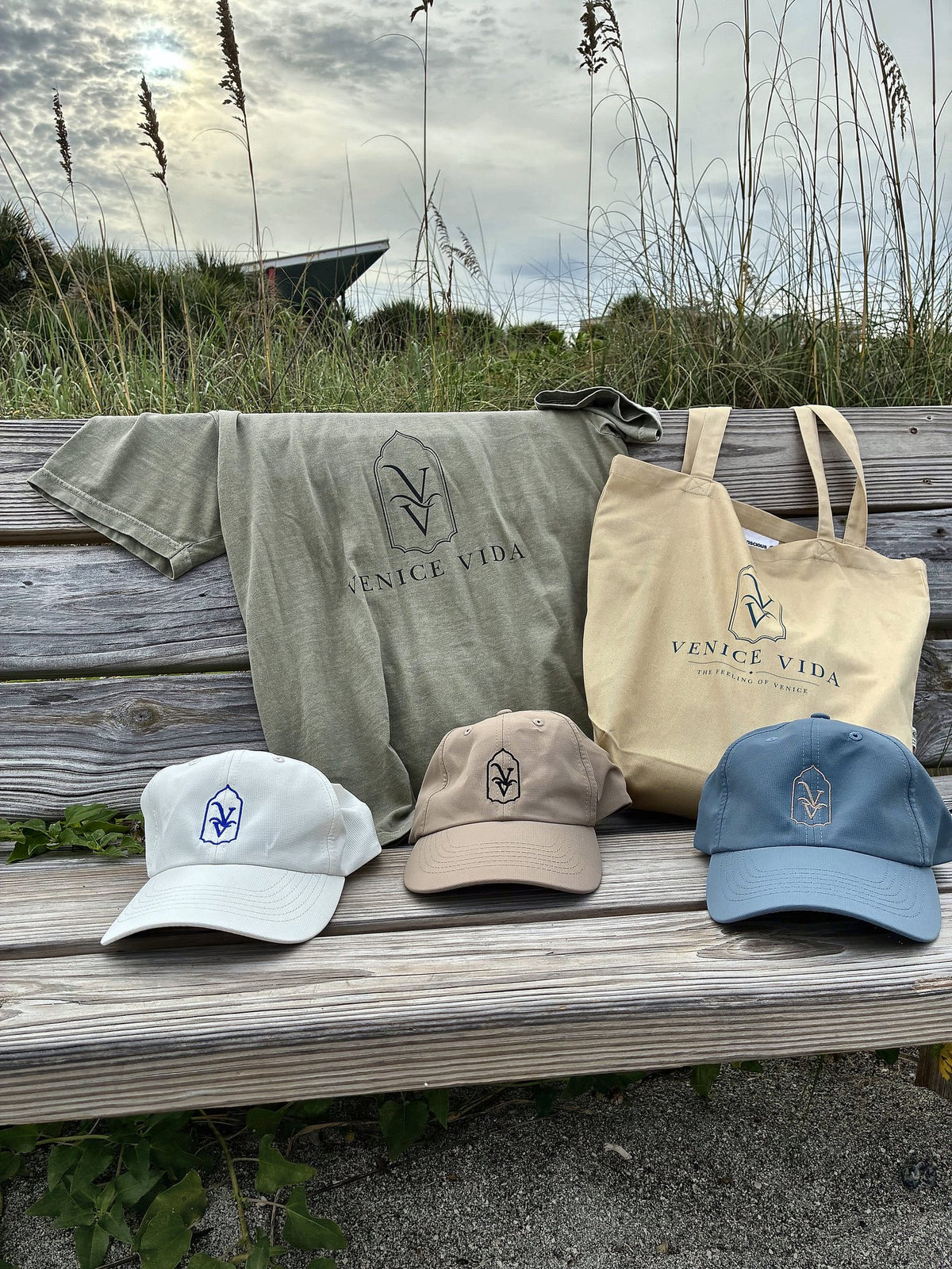 Venice Vida Coastal Neutral Statement Tee, Signature Market Tote and three VV hats on a beach boardwalk bench in Venice, Florida