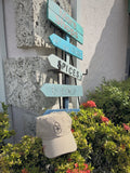 Venice Vida Heritage Hat in Khaki with black embroidered VV mark beside hand-painted shop signs in downtown Venice, Florida